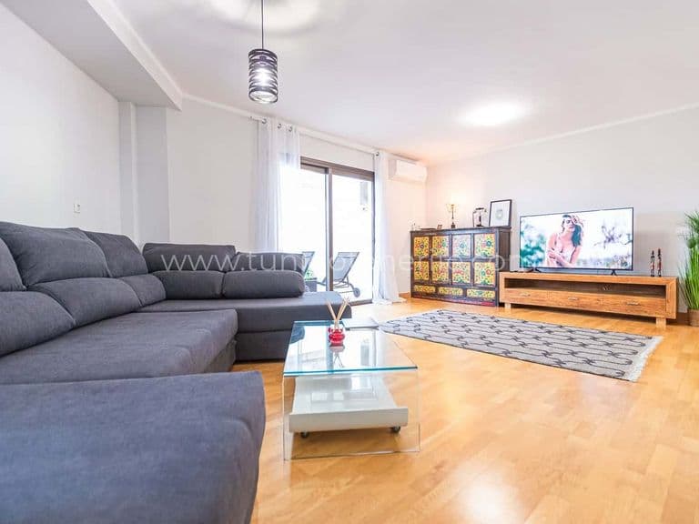 Salón luminoso con sofá gris, televisor y suelo de madera