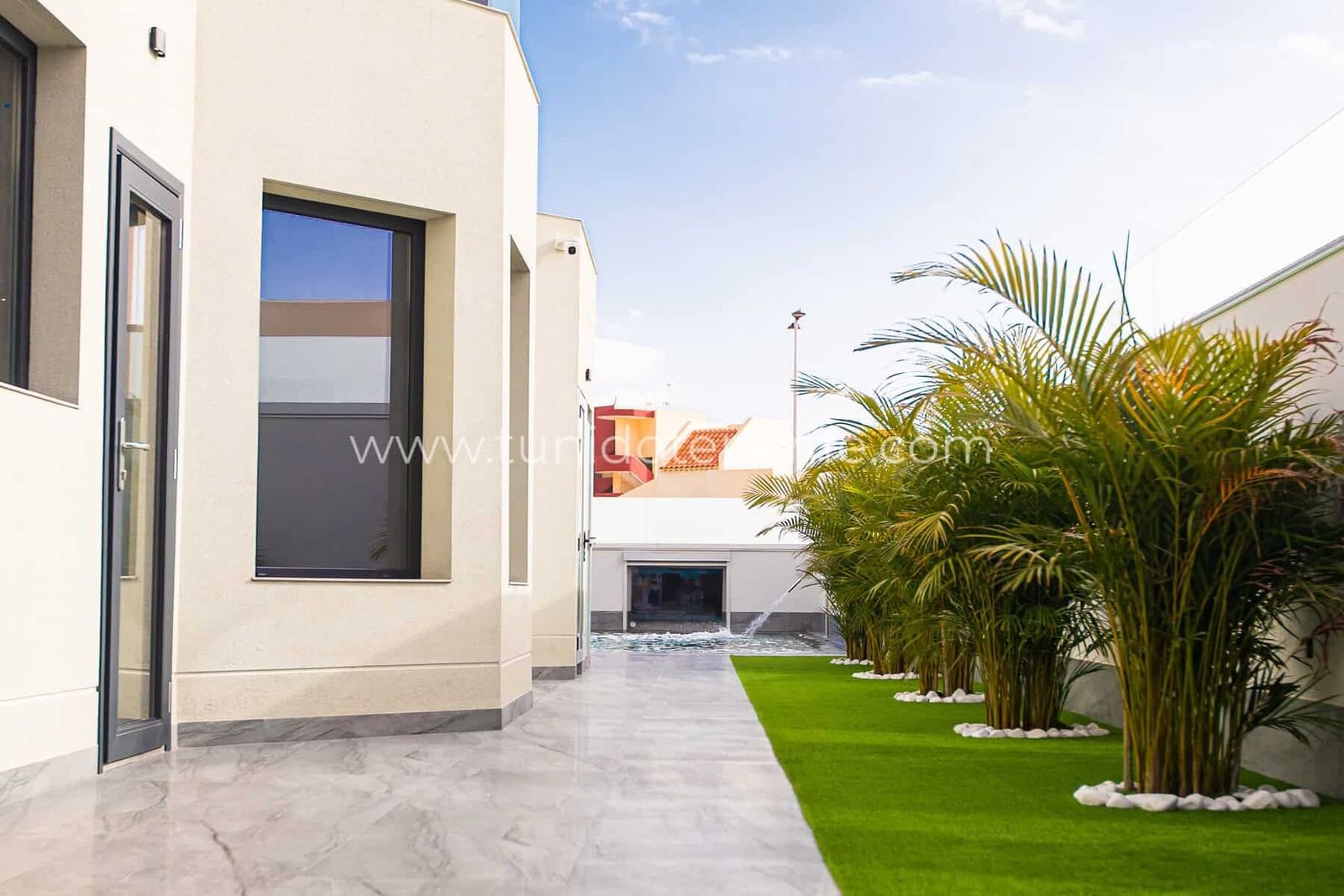 Patio exterior con suelo de piedra, césped artificial y palmeras