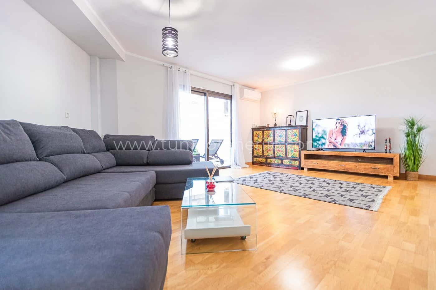 Salón luminoso con sofá gris, televisor y suelo de madera